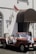 A young woman in a white long-sleeve shirt and black pants stands casually in front of a vintage red convertible car with open sides. The car is parked next to a building with white walls, adorned with an Indonesian flag and a brown awning. There's also a bicycle leaning against the wall.