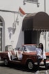 A young woman in a white long-sleeve shirt and black pants stands casually in front of a vintage red convertible car with open sides. The car is parked next to a building with white walls, adorned with an Indonesian flag and a brown awning. There's also a bicycle leaning against the wall.