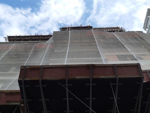 A construction site with scaffolding and a building partially covered in protective netting. The photo is taken from a low angle, focusing upwards towards the sky, which is partly cloudy. The building appears to be under renovation or construction, with structural beams visible at the bottom.