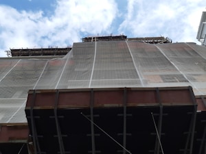 A construction site with scaffolding and a building partially covered in protective netting. The photo is taken from a low angle, focusing upwards towards the sky, which is partly cloudy. The building appears to be under renovation or construction, with structural beams visible at the bottom.