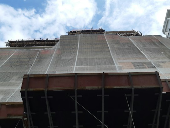 A construction site with scaffolding and a building partially covered in protective netting. The photo is taken from a low angle, focusing upwards towards the sky, which is partly cloudy. The building appears to be under renovation or construction, with structural beams visible at the bottom.