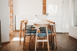 A minimalist dining area with a wooden table and eight chairs featuring light blue cushions. The room has a wooden floor and exposed brick elements on one wall, complemented by a radiator and a white door. Natural light illuminates the space, creating a warm and inviting atmosphere.
