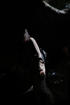 A person stands partially obscured in the shadows of what appears to be a natural setting, with light highlighting parts of the figure including their hair and shoulder. The dark clothing blends into the surrounding darkness, while subtle natural light creates a dramatic contrast.