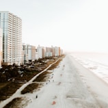 A coastal scene featuring a wide sandy beach stretching alongside a line of tall beachfront hotels and high-rise buildings. The beach is sparsely populated with people enjoying the sun and the waves. Soft waves roll onto the shore, and there are partially cloudy skies with a warm, hazy sunlight.