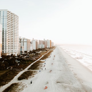 A coastal scene featuring a wide sandy beach stretching alongside a line of tall beachfront hotels and high-rise buildings. The beach is sparsely populated with people enjoying the sun and the waves. Soft waves roll onto the shore, and there are partially cloudy skies with a warm, hazy sunlight.