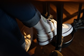 A person is wearing white socks with a colorful embroidered design along with white sneakers. The person is seated, and their legs are resting on a footrest under a table. The lighting creates a warm, cozy atmosphere.