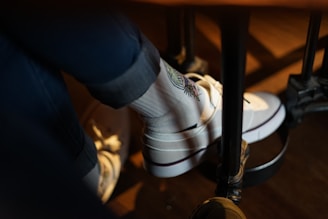 A person is wearing white socks with a colorful embroidered design along with white sneakers. The person is seated, and their legs are resting on a footrest under a table. The lighting creates a warm, cozy atmosphere.