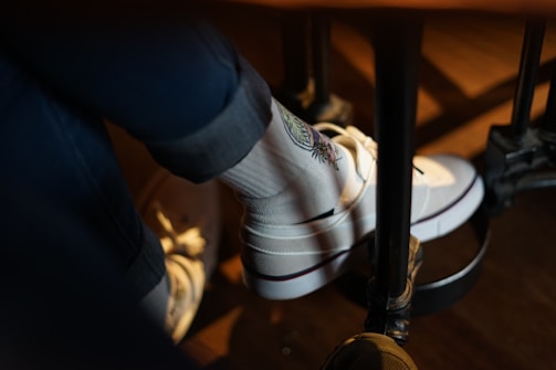 A person is wearing white socks with a colorful embroidered design along with white sneakers. The person is seated, and their legs are resting on a footrest under a table. The lighting creates a warm, cozy atmosphere.