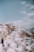 A picturesque landscape featuring traditional Cycladic architecture with whitewashed buildings and blue-domed roofs cascading down a hillside under a clear sky. The buildings are tightly packed, with narrow pathways winding between them, creating a stunning view.