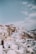 A picturesque landscape featuring traditional Cycladic architecture with whitewashed buildings and blue-domed roofs cascading down a hillside under a clear sky. The buildings are tightly packed, with narrow pathways winding between them, creating a stunning view.