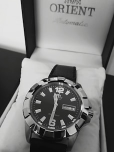 A close-up view of a stylish men's wristwatch with a black dial and bold silver markers. The watch is placed inside a white watch box featuring the brand name on the lid. It has a black leather strap and displays the day and date prominently.