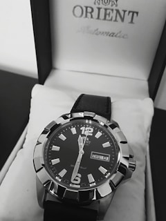 A close-up view of a stylish men's wristwatch with a black dial and bold silver markers. The watch is placed inside a white watch box featuring the brand name on the lid. It has a black leather strap and displays the day and date prominently.