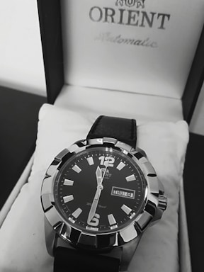 A close-up view of a stylish men's wristwatch with a black dial and bold silver markers. The watch is placed inside a white watch box featuring the brand name on the lid. It has a black leather strap and displays the day and date prominently.