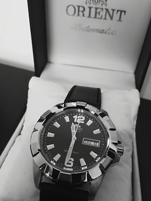 A close-up view of a stylish men's wristwatch with a black dial and bold silver markers. The watch is placed inside a white watch box featuring the brand name on the lid. It has a black leather strap and displays the day and date prominently.