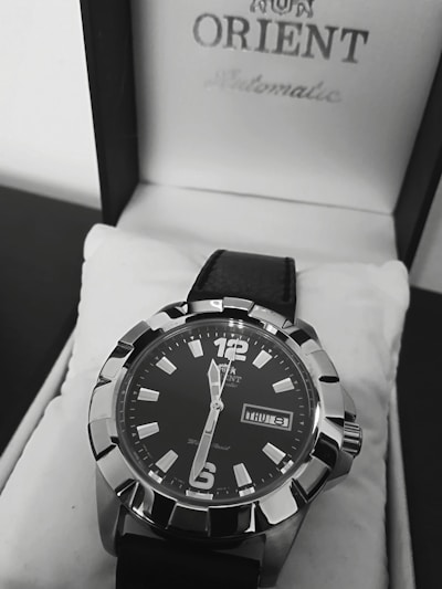 A close-up view of a stylish men's wristwatch with a black dial and bold silver markers. The watch is placed inside a white watch box featuring the brand name on the lid. It has a black leather strap and displays the day and date prominently.