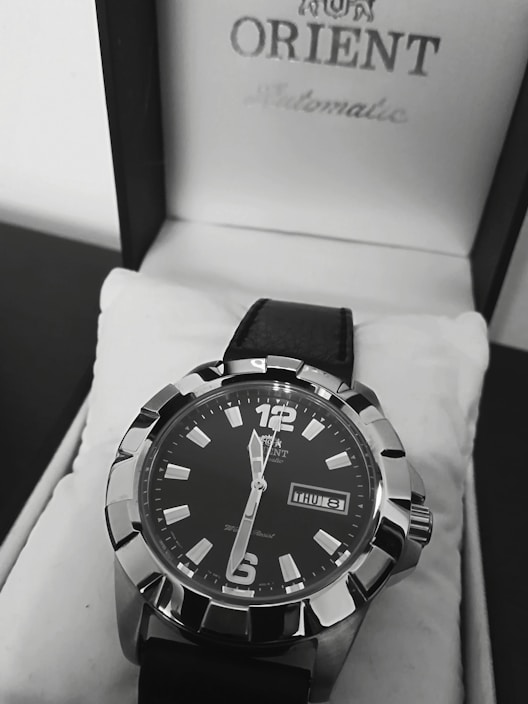 A close-up view of a stylish men's wristwatch with a black dial and bold silver markers. The watch is placed inside a white watch box featuring the brand name on the lid. It has a black leather strap and displays the day and date prominently.