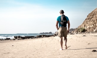 A person wearing a blue shirt and khaki shorts is walking barefoot on a sandy beach. They have a backpack and are facing away, looking towards the ocean. There is a rocky cliff to the right and waves gently crashing against the shore.