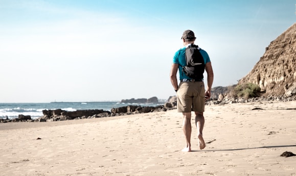 A person wearing a blue shirt and khaki shorts is walking barefoot on a sandy beach. They have a backpack and are facing away, looking towards the ocean. There is a rocky cliff to the right and waves gently crashing against the shore.