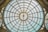A large, intricate glass dome ceiling featuring a geometric pattern with radiating lines extending from the center. The structure is made of metal and glass panels, creating a vast and detailed architectural design.