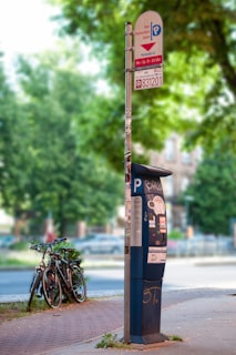 A parking meter stands on a sidewalk next to a sign indicating parking information, surrounded by trees and greenery. There is some graffiti on the meter, and a few bicycles are parked nearby.