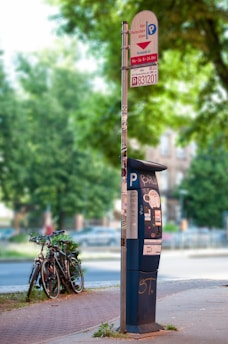 A parking meter stands on a sidewalk next to a sign indicating parking information, surrounded by trees and greenery. There is some graffiti on the meter, and a few bicycles are parked nearby.