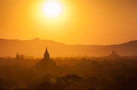 A breathtaking sunset casts a golden hue over an expansive landscape filled with ancient pagodas. The silhouette of these structures is highlighted against the bright, descending sun, creating a serene and mystical atmosphere. The distant mountain range adds depth to the scene, completing the picturesque view.