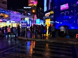 A lively urban street scene at night featuring colorful neon lights and various shops with bright signage. People are walking and socializing on the wet pavement, reflecting the vibrant lights. A street sign indicates the location as Church Street, and the atmosphere is bustling with activity.