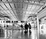 A busy airport terminal with a high, metallic ceiling and multiple travelers walking through. Two people are seen from behind pulling rolling luggage. Signs and gates are visible in the background, indicating areas such as A49.