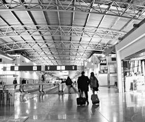 A busy airport terminal with a high, metallic ceiling and multiple travelers walking through. Two people are seen from behind pulling rolling luggage. Signs and gates are visible in the background, indicating areas such as A49.