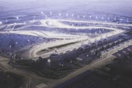 An aerial view of a well-lit, winding racetrack surrounded by desert terrain. Several brightly illuminated track lights highlight the curving paths. The area includes multiple structures such as grandstands or pavilions, with organized rows of them to the side.