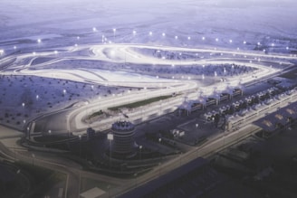 An aerial view of a well-lit, winding racetrack surrounded by desert terrain. Several brightly illuminated track lights highlight the curving paths. The area includes multiple structures such as grandstands or pavilions, with organized rows of them to the side.