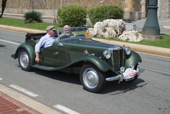 A vintage green convertible car is driving on a road with two people seated inside. The car has a retro design with chrome details and a classic grille. It appears to be part of an event, indicated by a sign with the number 35 attached to the front. The background includes a stone wall, greenery, and a lamp post.