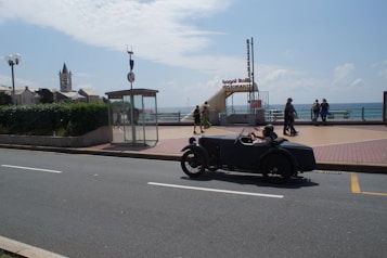 A classic car with two passengers drives along a road by the sea. In the background, there is a building with a tower, a public telephone booth, and a restaurant entrance labeled 'bagni italiani'. Several people are walking near the seaside promenade, enjoying a sunny day. The ocean is visible under a partly cloudy sky.