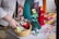 A close-up view of a person arranging a selection of handmade dolls on a table. The dolls are dressed in colorful traditional costumes with intricate patterns. In the foreground, there is a basket filled with stuffed toys. The background shows a variety of packaged items, possibly crafted objects.