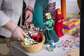 A close-up view of a person arranging a selection of handmade dolls on a table. The dolls are dressed in colorful traditional costumes with intricate patterns. In the foreground, there is a basket filled with stuffed toys. The background shows a variety of packaged items, possibly crafted objects.