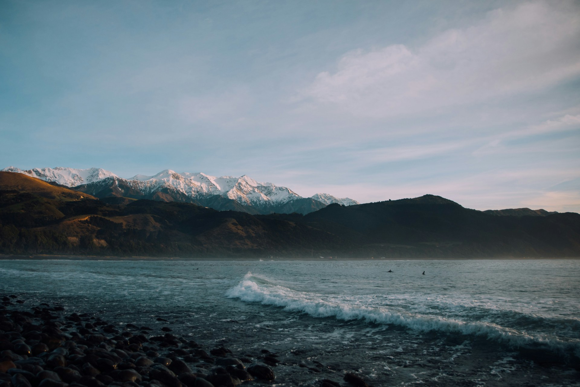 Snow-capped mountains rise majestically in the distance, with dark, forested hills below. A calm sea stretches out in the foreground, waves gently lapping against a rocky shoreline. The scene is illuminated by soft, natural light under a partially cloudy sky.
