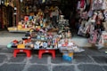 An array of colorful souvenirs, trinkets, and handmade crafts is displayed outside a shop. The items include decorative masks, miniature sculptures, bracelets, postcards, and patterned handbags. The setup is vibrant and visually engaging, with a diverse range of items closely packed on small tables and hanging on the shop walls.