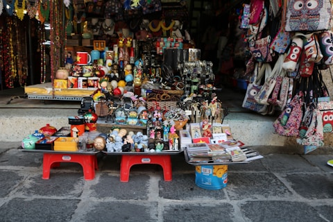 An array of colorful souvenirs, trinkets, and handmade crafts is displayed outside a shop. The items include decorative masks, miniature sculptures, bracelets, postcards, and patterned handbags. The setup is vibrant and visually engaging, with a diverse range of items closely packed on small tables and hanging on the shop walls.
