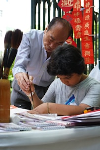 An elderly man is instructing a young boy in the art of calligraphy. They are both focused, with the older man guiding the boy's hand as he writes with a brush. Red banners with Chinese characters are visible in the background, adding cultural context to the scene.