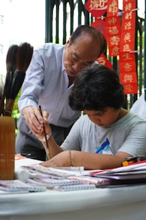 An elderly man is instructing a young boy in the art of calligraphy. They are both focused, with the older man guiding the boy's hand as he writes with a brush. Red banners with Chinese characters are visible in the background, adding cultural context to the scene.