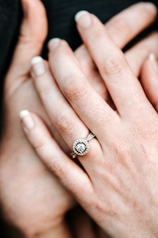 A set of hands with neatly manicured nails is shown with a sparkling engagement ring featuring a central diamond surrounded by smaller diamonds. The focus is on the elegance and detail of the jewelry, highlighting its craftsmanship and brilliance.