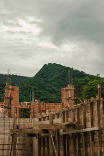 Construction site with partially built brick walls and wooden scaffolding, set against a backdrop of lush green mountains under a cloudy sky.