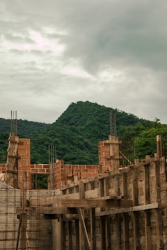Construction site with partially built brick walls and wooden scaffolding, set against a backdrop of lush green mountains under a cloudy sky.