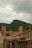 Construction site with partially built brick walls and wooden scaffolding, set against a backdrop of lush green mountains under a cloudy sky.