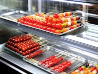 Assorted skewers of colorful candied fruits are neatly arranged on metal trays inside a glass display case. The fruits include vibrant red cherry tomatoes and various other fruits in shades of orange, yellow, and green. The glossy sheen of the candy coating gives a fresh and appealing look.
