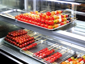 Assorted skewers of colorful candied fruits are neatly arranged on metal trays inside a glass display case. The fruits include vibrant red cherry tomatoes and various other fruits in shades of orange, yellow, and green. The glossy sheen of the candy coating gives a fresh and appealing look.