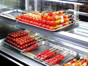 Assorted skewers of colorful candied fruits are neatly arranged on metal trays inside a glass display case. The fruits include vibrant red cherry tomatoes and various other fruits in shades of orange, yellow, and green. The glossy sheen of the candy coating gives a fresh and appealing look.