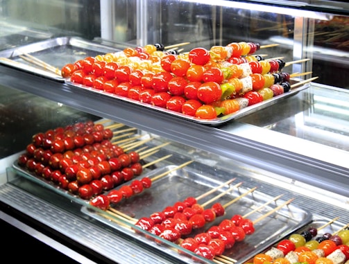 Assorted skewers of colorful candied fruits are neatly arranged on metal trays inside a glass display case. The fruits include vibrant red cherry tomatoes and various other fruits in shades of orange, yellow, and green. The glossy sheen of the candy coating gives a fresh and appealing look.