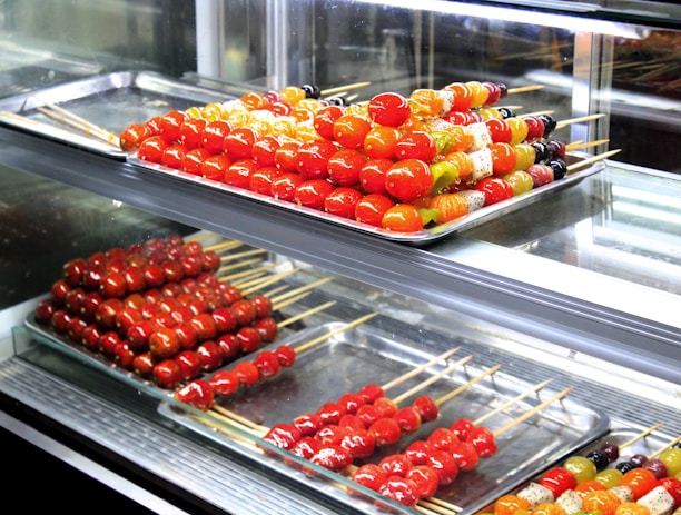 Assorted skewers of colorful candied fruits are neatly arranged on metal trays inside a glass display case. The fruits include vibrant red cherry tomatoes and various other fruits in shades of orange, yellow, and green. The glossy sheen of the candy coating gives a fresh and appealing look.