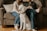 Two people are sitting on a couch, one holding a cat, while a large white dog is sitting in front of them. The setting appears cozy with pillows and a wooden floor visible. The mood suggests a relaxed, domestic atmosphere.